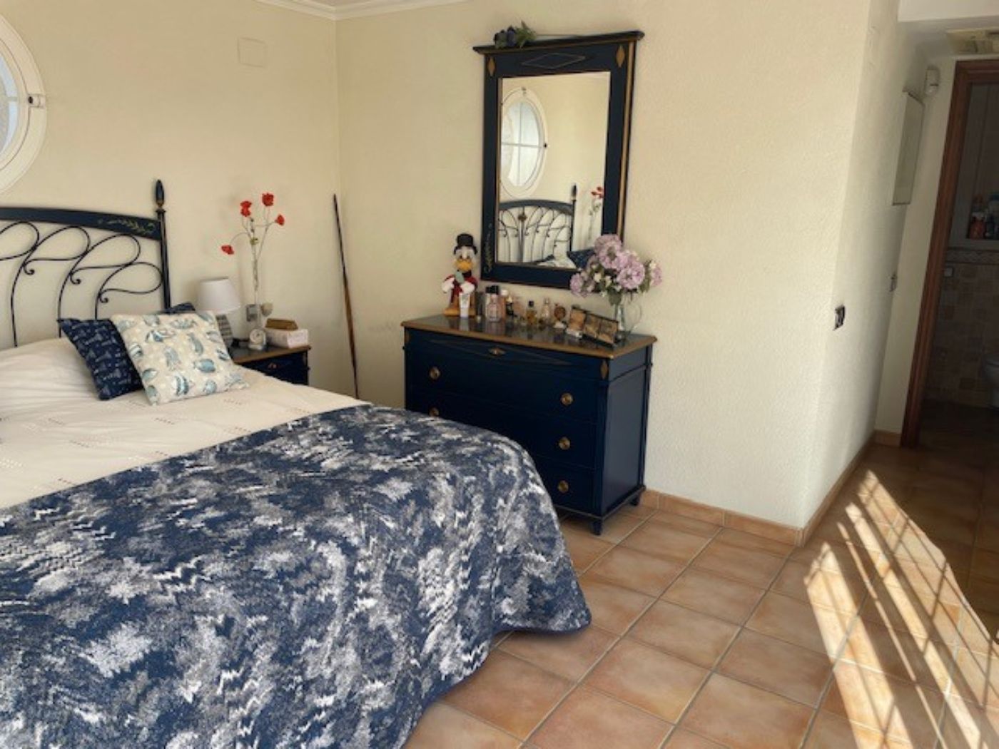 Chambre avec tête de lit en fer forgé, commode bleu marine avec miroir assorti et sol en terre cuite. Une fenêtre circulaire apporte une lumière naturelle.