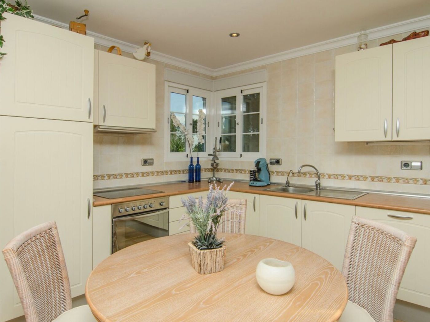 Cuisine avec placards crème, plans de travail en bois et coin repas comprenant une table ronde et des chaises en osier sous une fenêtre d'angle.