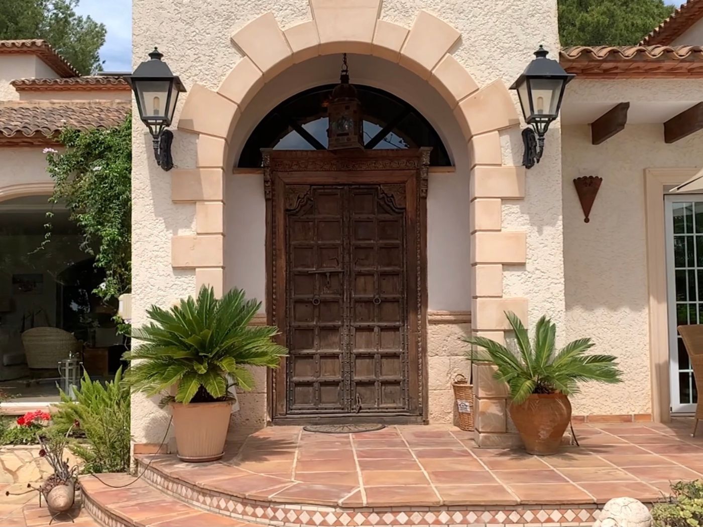 Entrée avec double porte en bois sculpté ancien sous une arche en pierre, encadrée de lanternes noires et de palmiers cycas sur un porche en terre cuite.