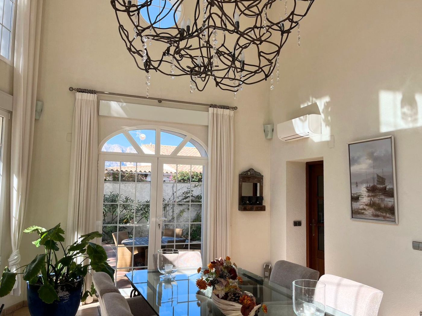 Espace salle à manger à double hauteur sous plafond avec lustre en fer forgé. Les portes-fenêtres cintrées offrent un accès direct à la terrasse.