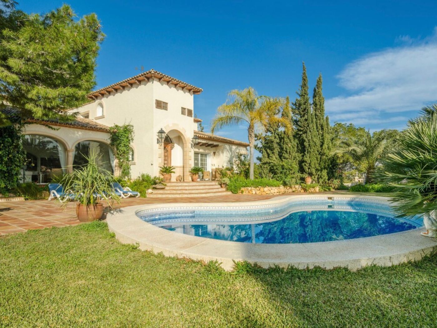 Extérieur de villa méditerranéenne avec piscine courbe, terrasse en terre cuite et façade en stuc blanc ornée d'arches et d'un jardin paysager arboré.