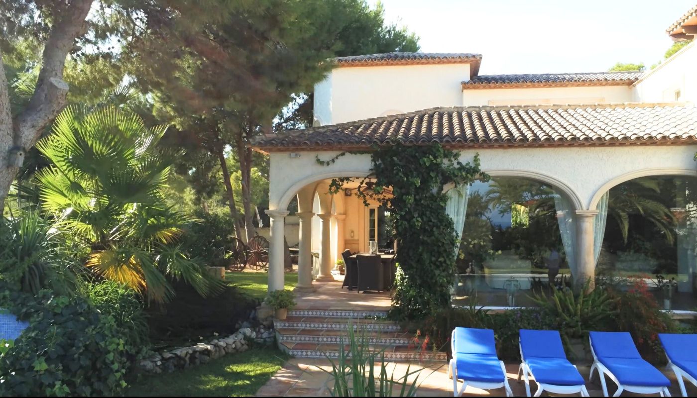 Extérieur d'une villa méditerranéenne avec porche à arcades, colonnes en pierre, toit en tuiles et terrasse équipée de bains de soleil bleus.