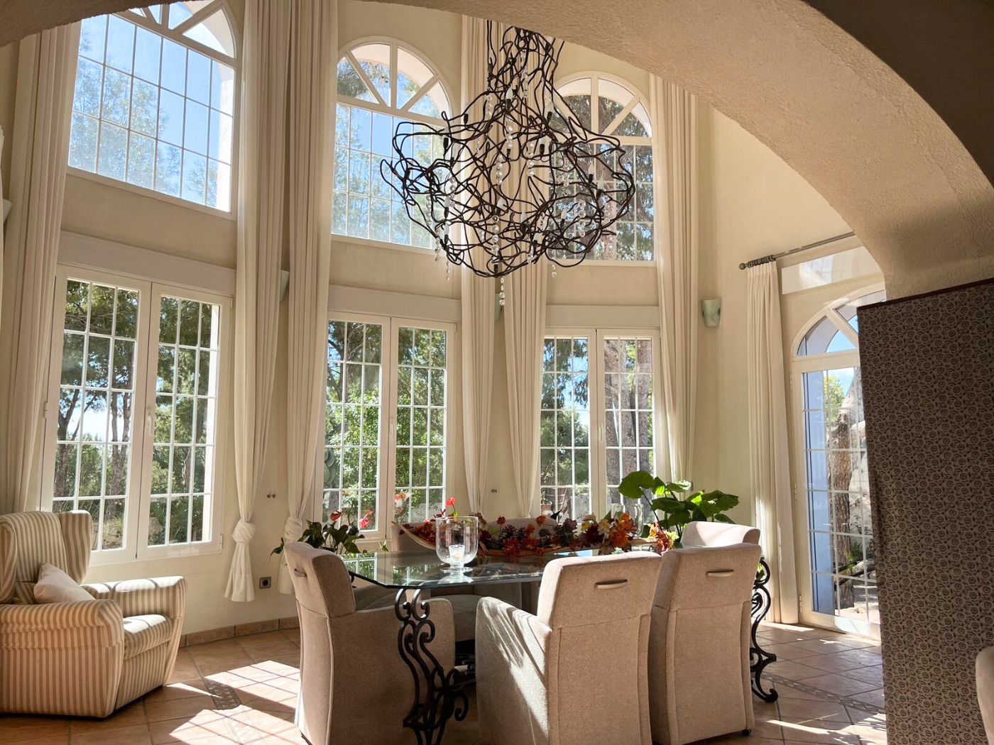 Salle à manger lumineuse avec plafonds double hauteur, fenêtres cintrées, table en verre sur pieds en fer forgé et lustre sculptural en fil de fer.