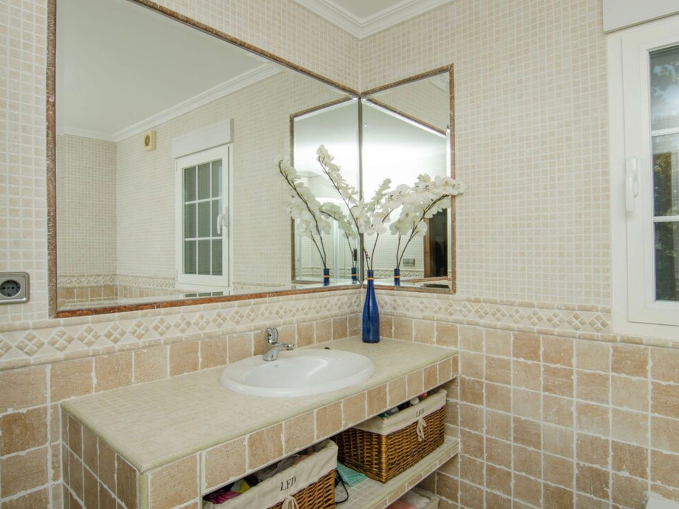 Salle de bain avec plan de toilette en carrelage beige, vasque intégrée et rangements en osier. Les miroirs d'angle reflètent les murs en mosaïque.