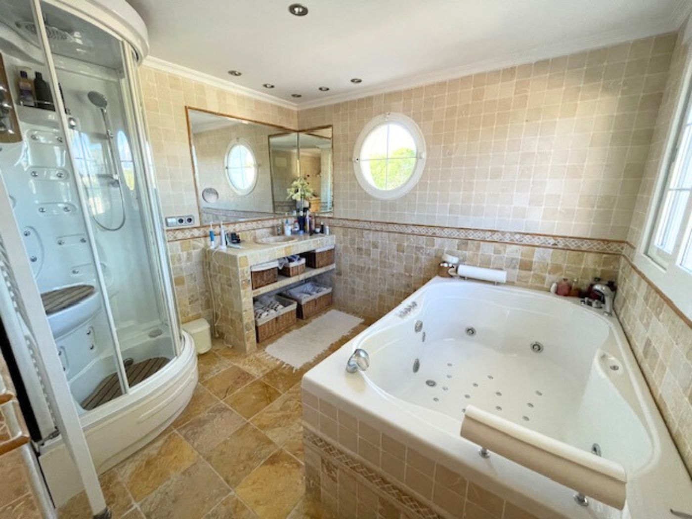 Salle de bain comprenant une baignoire d'hydromassage, une cabine de douche vitrée et des murs carrelés avec une fenêtre œil-de-boeuf.