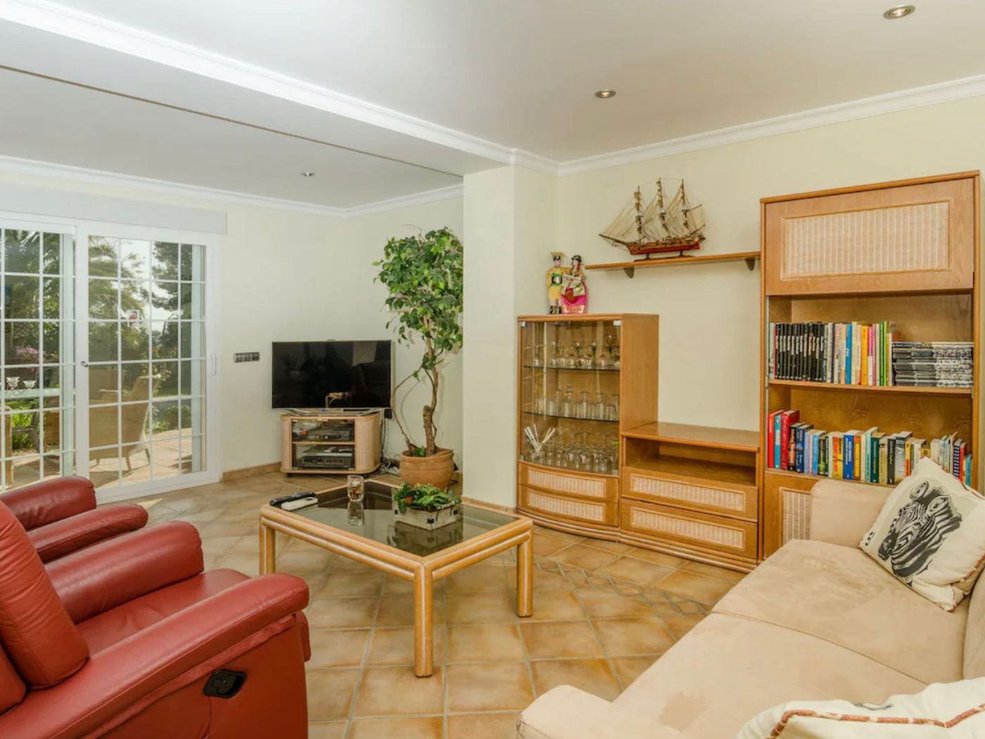 Salon avec sol en terre cuite, fauteuils en cuir rouge et mobilier en bois. Les baies vitrées coulissantes offrent une luminosité naturelle et un accès extérieur.
