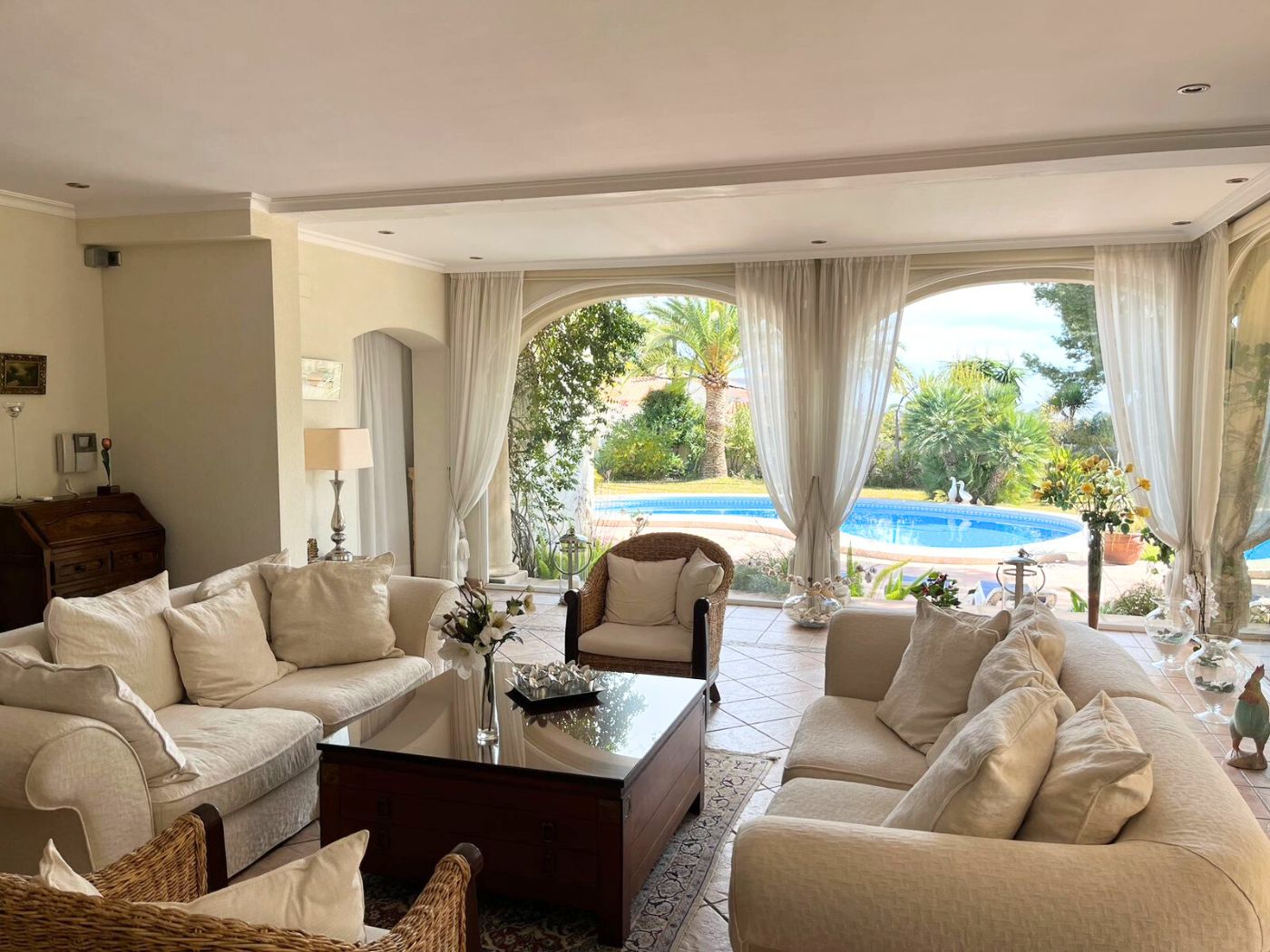 Salon spacieux avec canapés crème et fauteuils en osier, doté de grandes baies vitrées cintrées offrant une vue sur la piscine et le jardin arboré.
