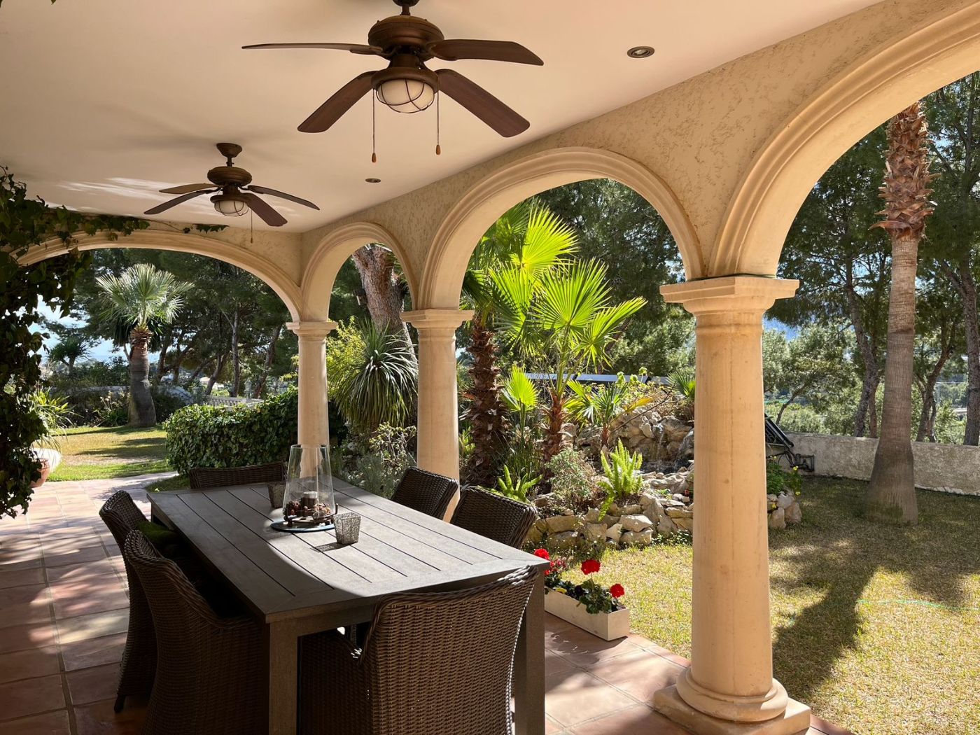 Terrasse couverte avec colonnade en pierre, sol en terre cuite et espace dînatoire extérieur offrant une vue sur un jardin méditerranéen arboré.