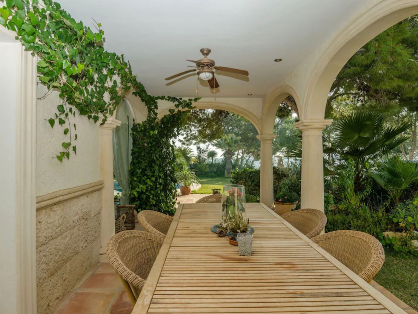 Terrasse couverte dotée d'arches en pierre, d'une table à manger en bois et de fauteuils en osier, avec vue sur un jardin arboré et ventilateur de plafond.