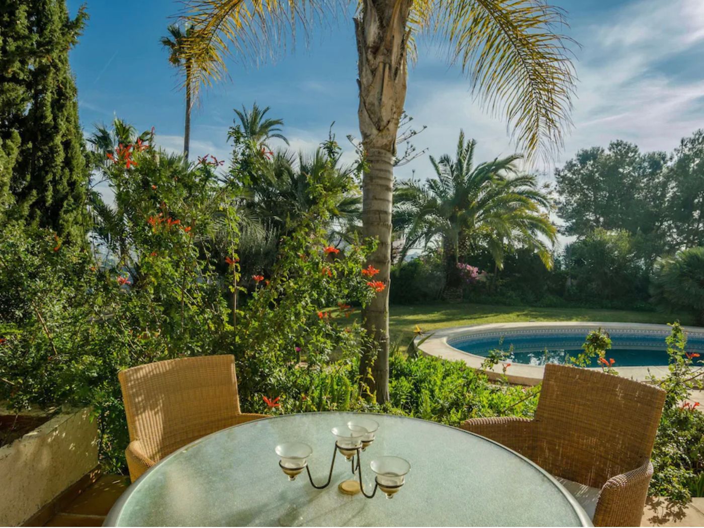 Terrasse extérieure avec table en verre et sièges en osier, offrant une vue sur une piscine circulaire et un jardin méditerranéen aux palmiers matures.