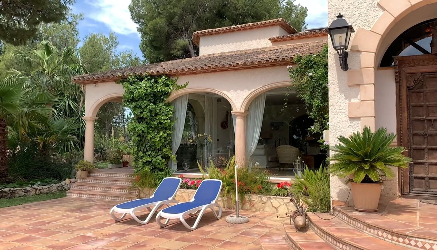 Terrasse extérieure en terre cuite avec chaises longues bleues, porche à arcades végétalisé et porte d'entrée monumentale en bois sculpté.