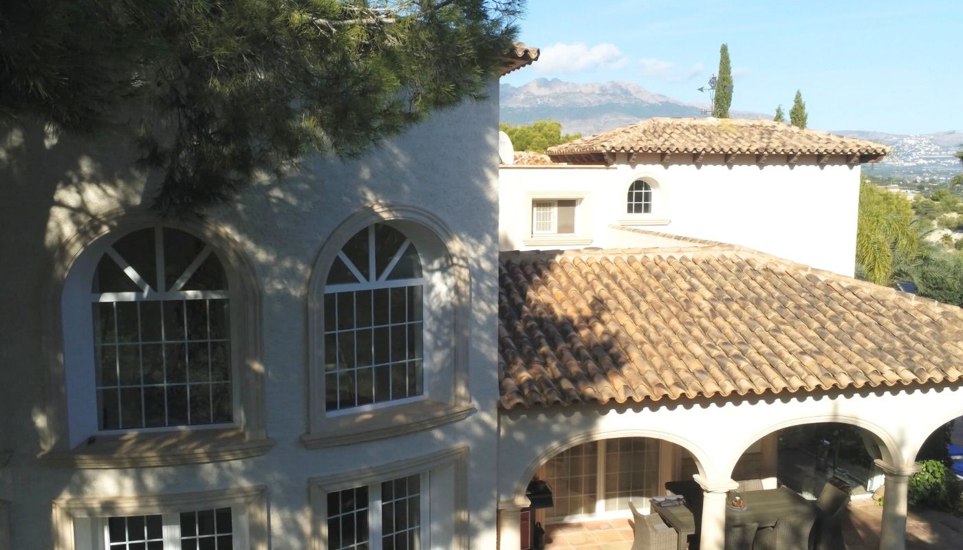 Vue extérieure d'une villa méditerranéenne avec toits en tuiles, fenêtres cintrées et terrasse couverte offrant une vue sur les montagnes.