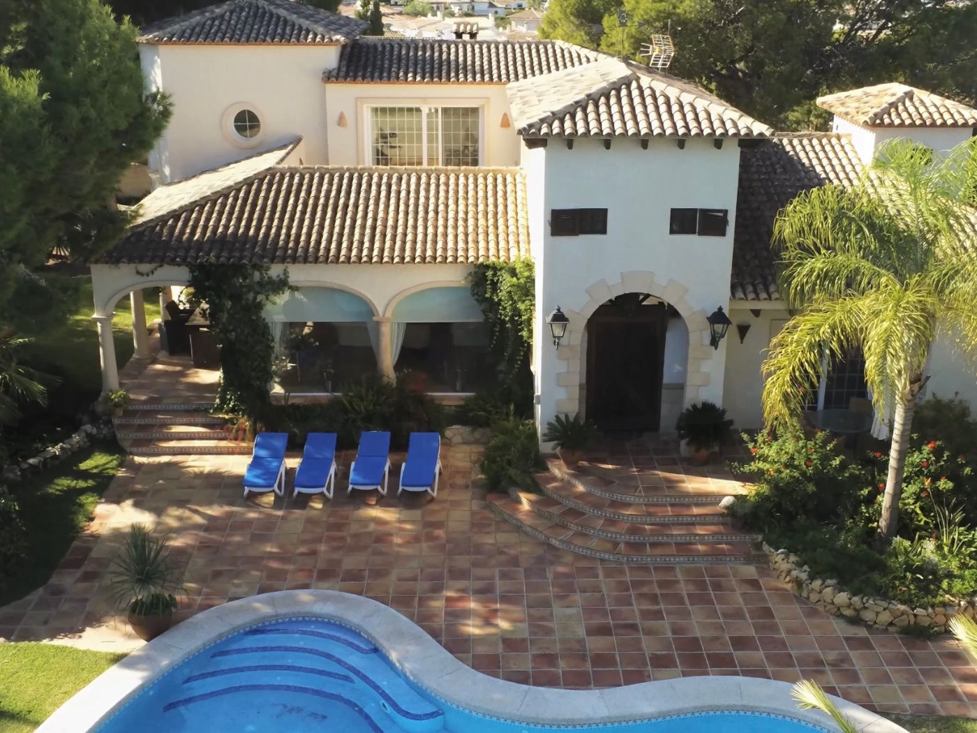 Vue surélevée d'une villa méditerranéenne avec piscine incurvée, terrasse en terre cuite avec quatre transats et façade blanche aux entrées voûtées.