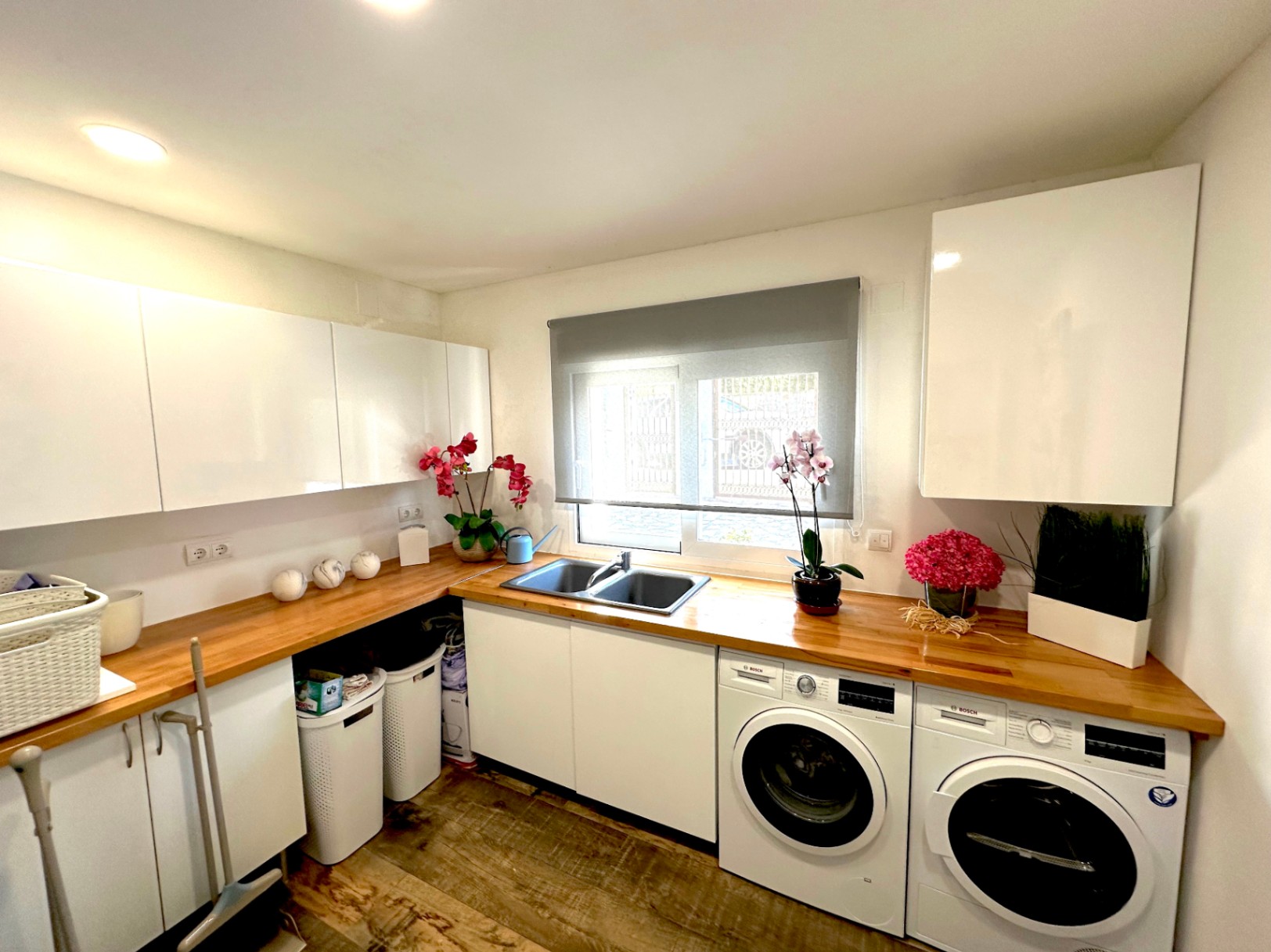 Buanderie fonctionnelle avec plan de travail en bois naturel, meubles blancs laqués et appareils Bosch intégrés sous une fenêtre avec store.