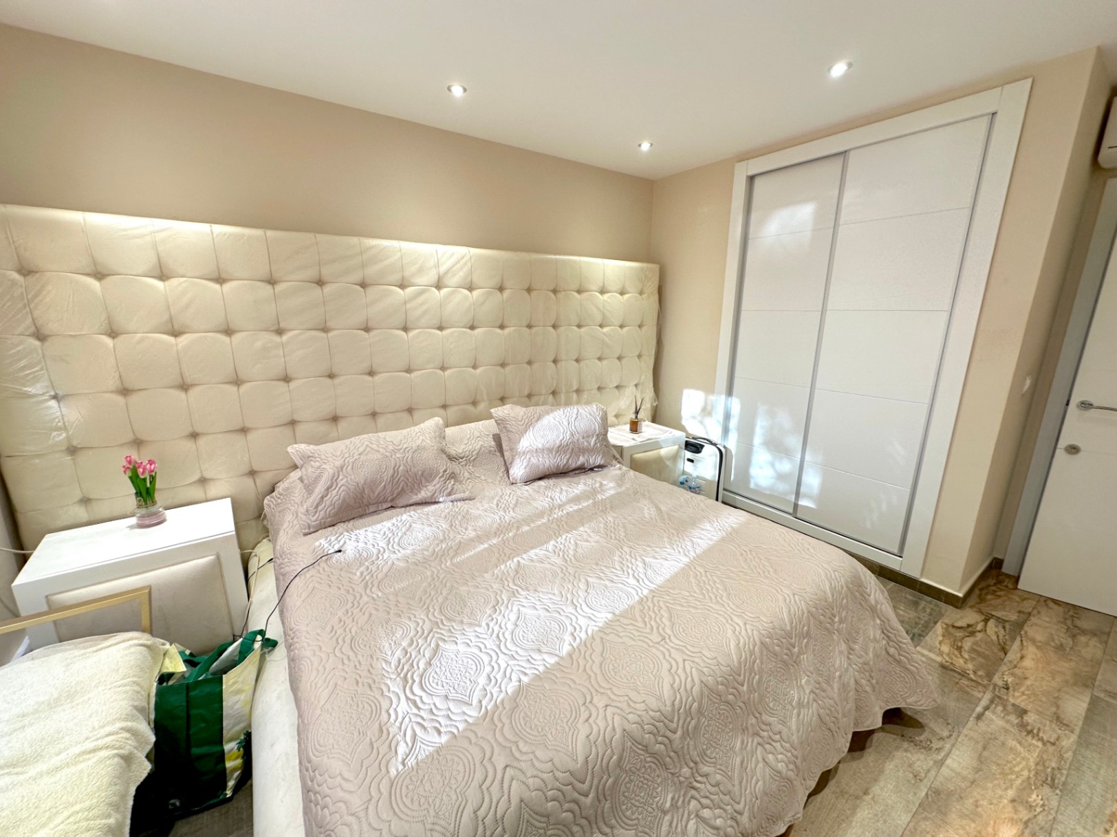 Chambre avec une grande tête de lit capitonnée, éclairage encastré au plafond et une armoire encastrée à portes coulissantes blanches sur un sol en pierre.