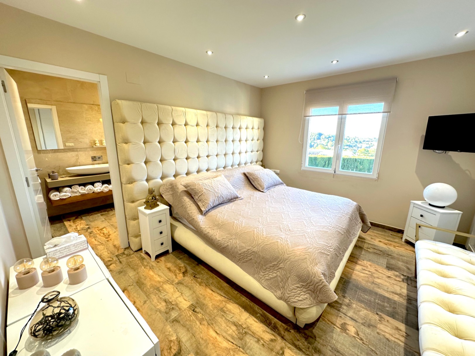 Chambre principale avec une grande tête de lit capitonnée, sol en grès cérame imitation bois et salle de bain attenante avec vasque à poser.