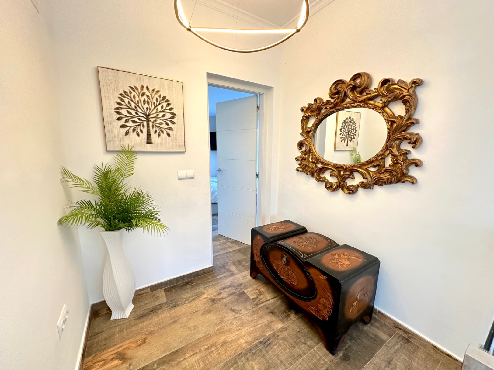 Couloir avec sol imitation bois, coffre sculpté classique et miroir doré ornemental, éclairé par un luminaire LED circulaire de design contemporain.