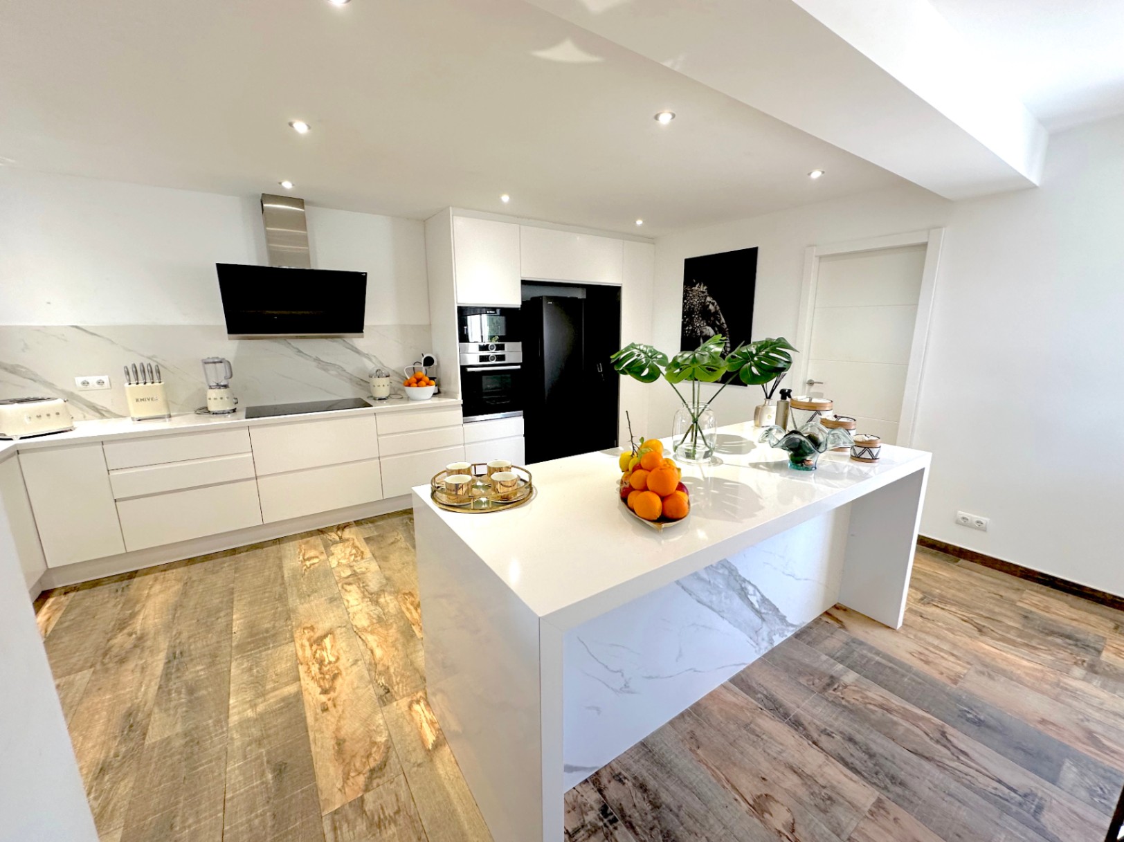 Cuisine design contemporaine avec îlot central en cascade effet marbre, meubles blancs minimalistes, électroménager noir intégré et sol en grès cérame imitation bois.