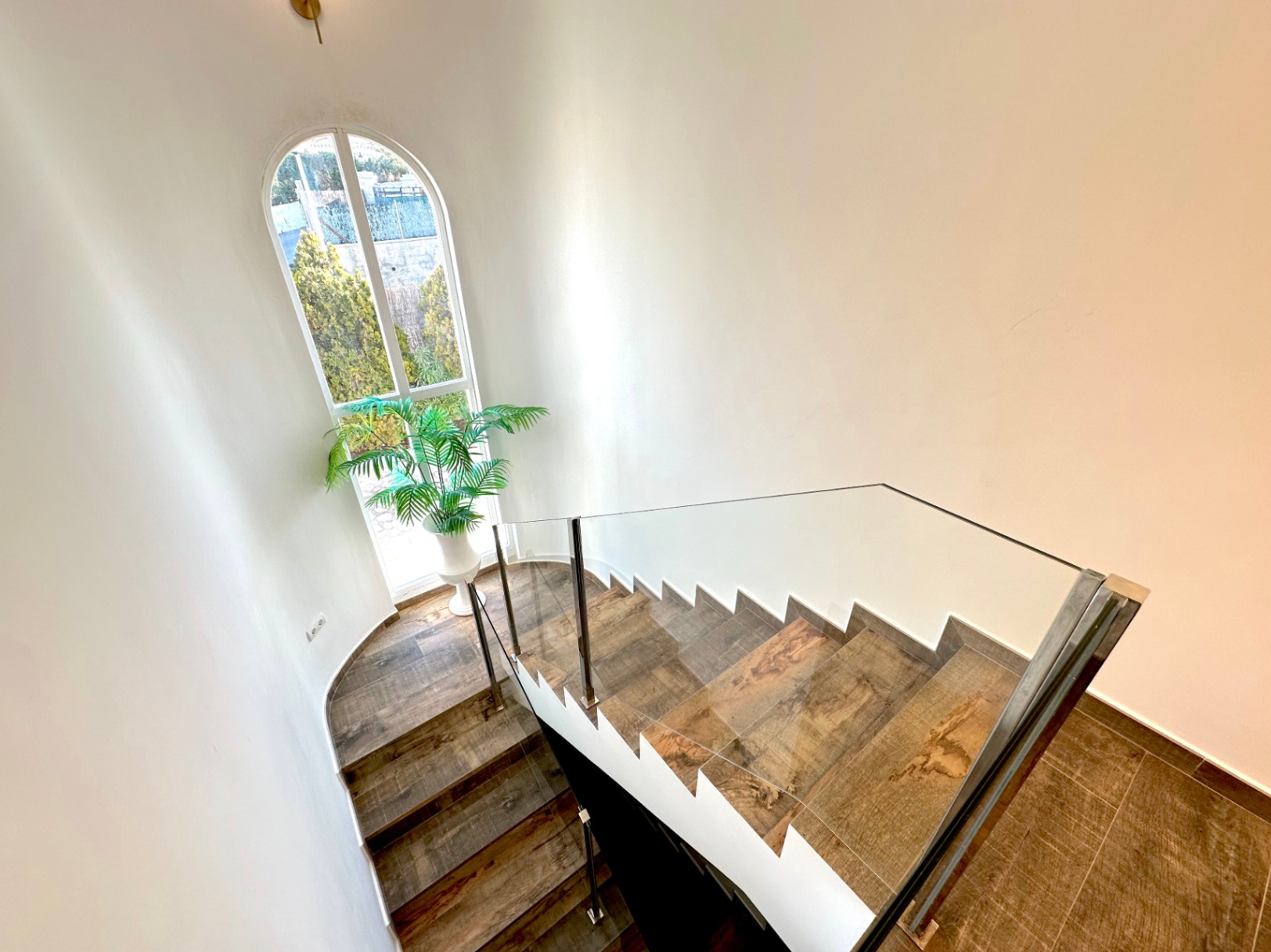 Escalier aux marches effet bois avec garde-corps en verre, éclairé par une grande fenêtre en plein cintre donnant sur l'extérieur verdoyant.