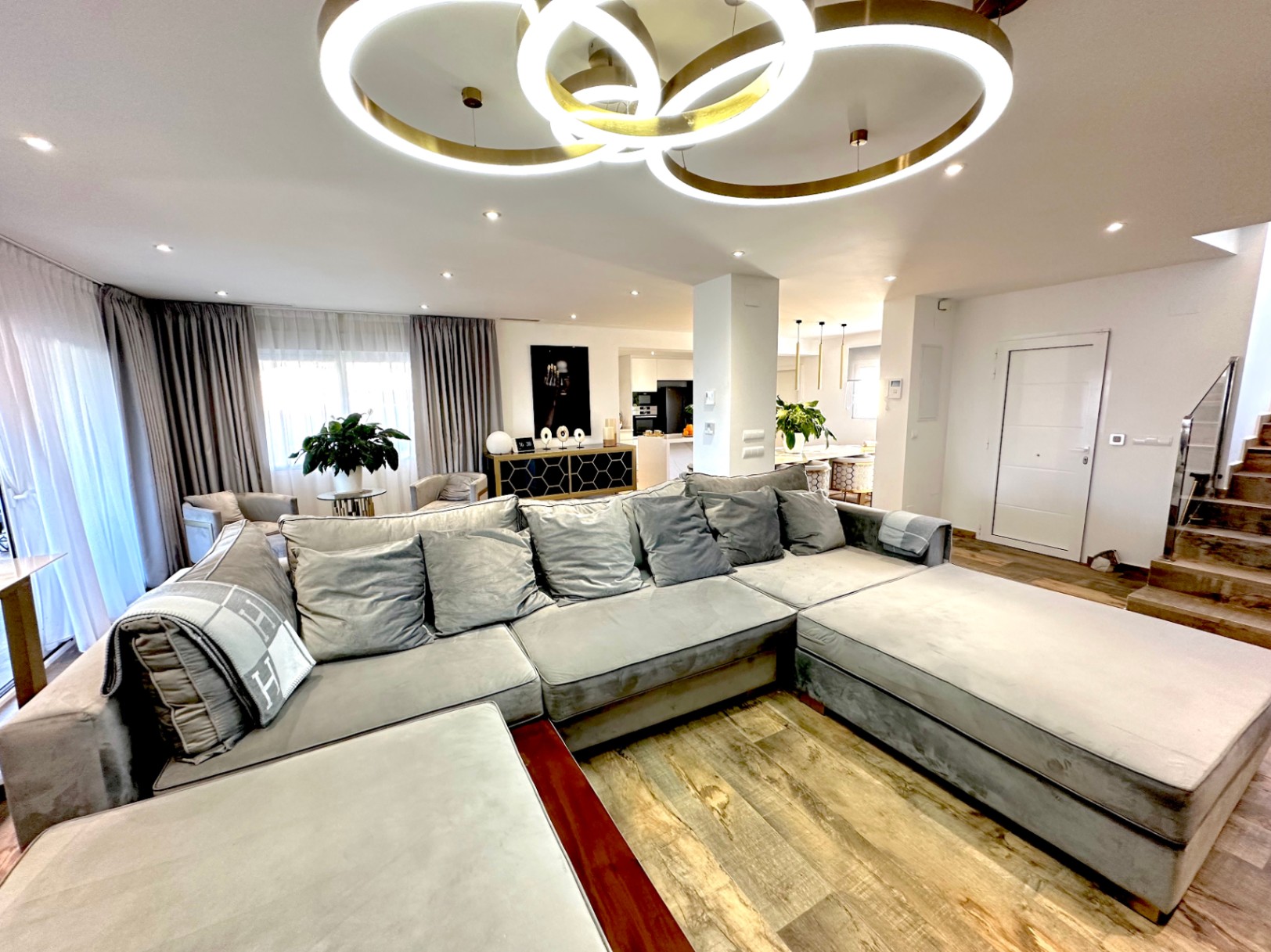 Espace de vie ouvert comprenant un grand canapé d'angle gris, un sol aspect bois et un luminaire circulaire moderne à LED sous un plafond à spots encastrés.