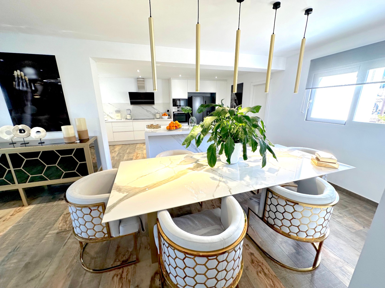 Salle à manger à aire ouverte avec table en marbre, chaises design aux détails dorés et luminaires suspendus, intégrée à une cuisine moderne équipée.