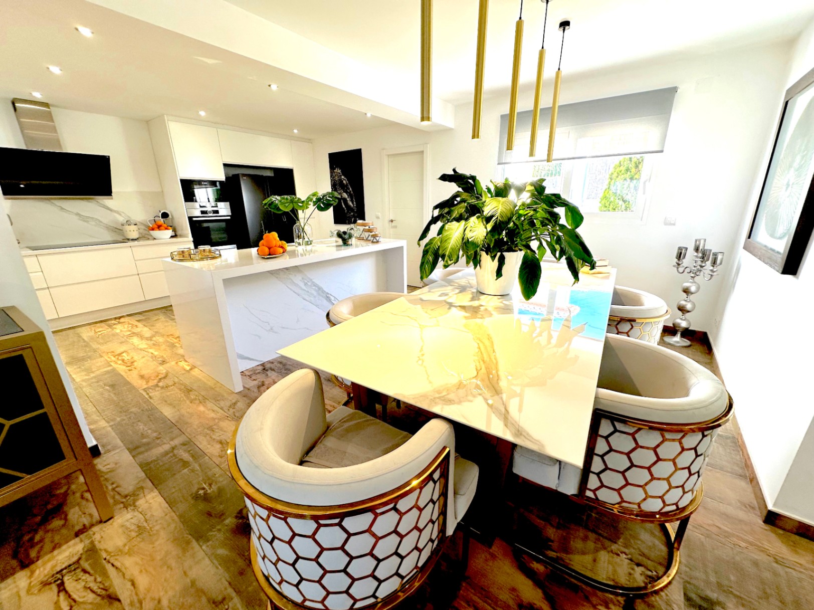 Salle à manger ouverte avec table en marbre et chaises aux détails dorés, communiquant avec une cuisine moderne dotée d'un îlot central et de luminaires suspendus.