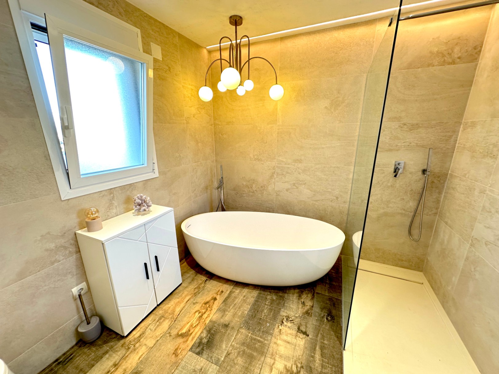 Salle de bain avec baignoire îlot ovale, douche à l'italienne vitrée, sol en grès cérame aspect bois et éclairage design avec lustre à globes.