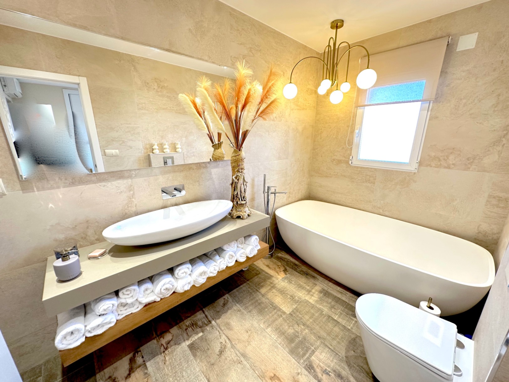 Salle de bains moderne avec baignoire îlot, vasque à poser et WC suspendu. Elle dispose d'un lustre design doré et de carrelage mural aux tons sable.