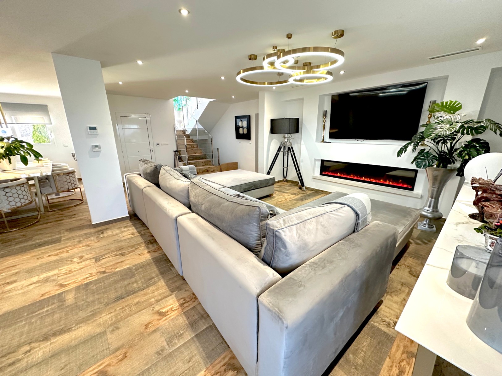 Salon à aire ouverte avec grand canapé gris, cheminée linéaire intégrée sous le téléviseur et luminaires circulaires. Sol en grès imitation bois et escalier avec garde-corps en verre.