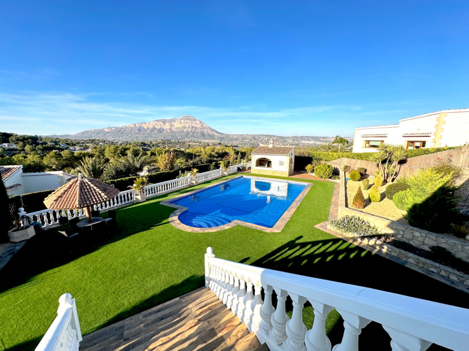 Vue en surplomb de la piscine entourée de gazon synthétique, avec balustrade blanche, cuisine d'été et panorama sur la montagne Montgó.