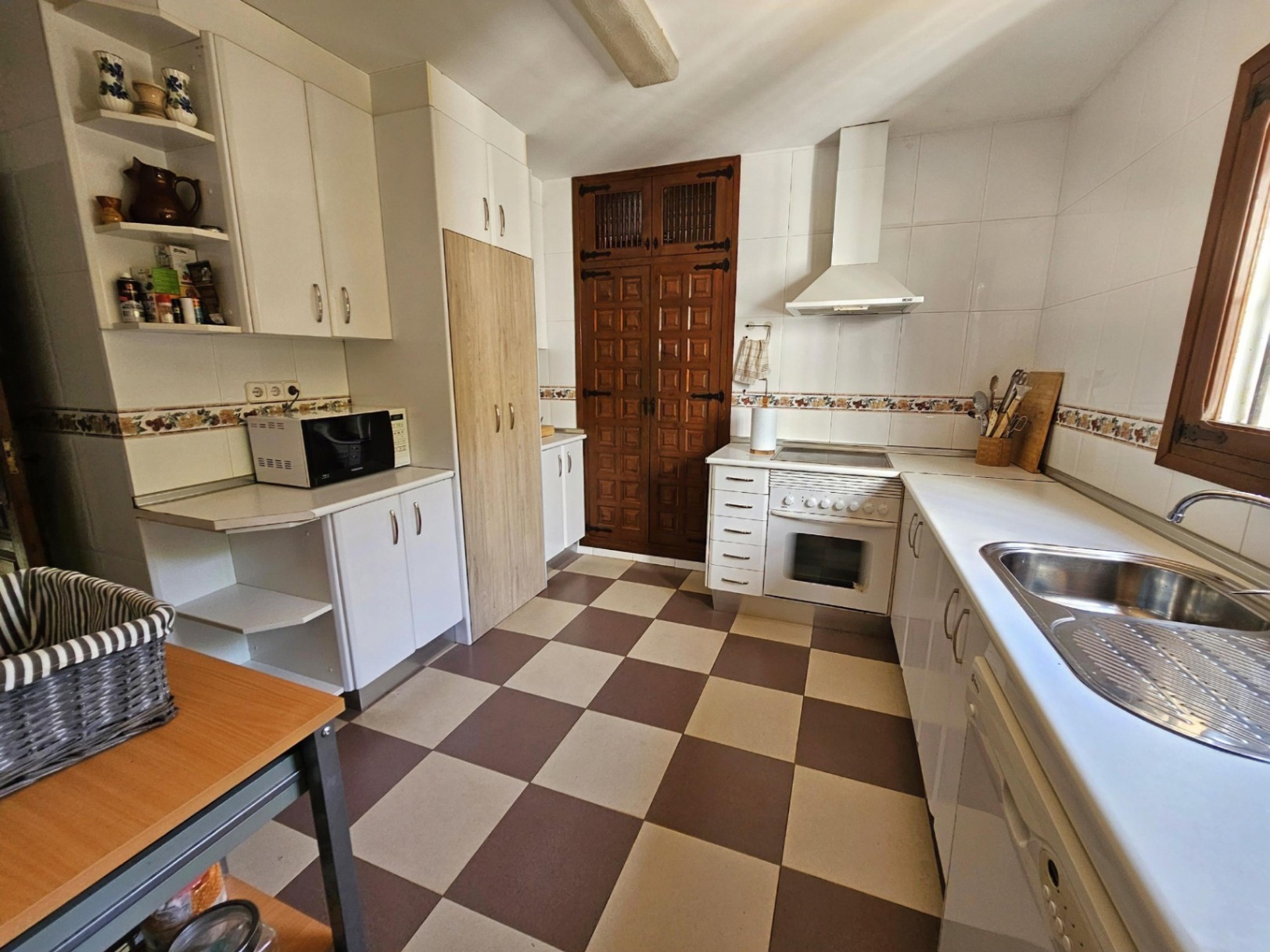 Cuisine avec carrelage en damier, meubles blancs et une porte de cellier en bois rustique sculpté. Équipée d'un plan de travail blanc et d'un évier en inox.