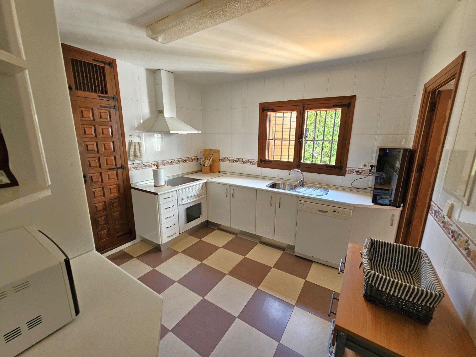 Cuisine avec placards blancs, sol en damier bicolore et finitions en bois sombre. Équipée d'un four blanc et d'un lave-vaisselle sous un plan de travail clair.