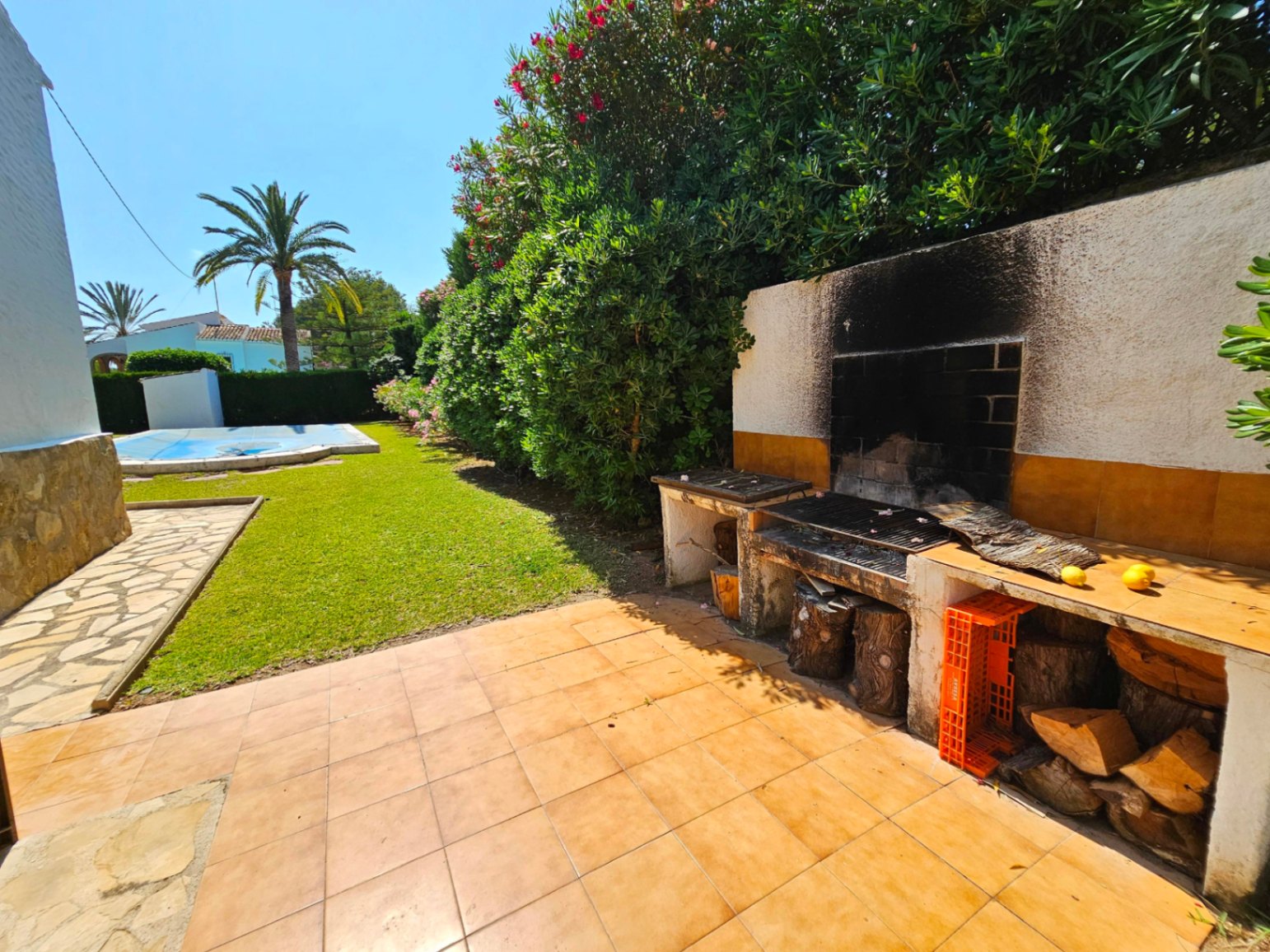 Espace barbecue maçonné avec plans de travail en carrelage et rangement pour le bois, bordant une pelouse soignée et une piscine entourée de palmiers.