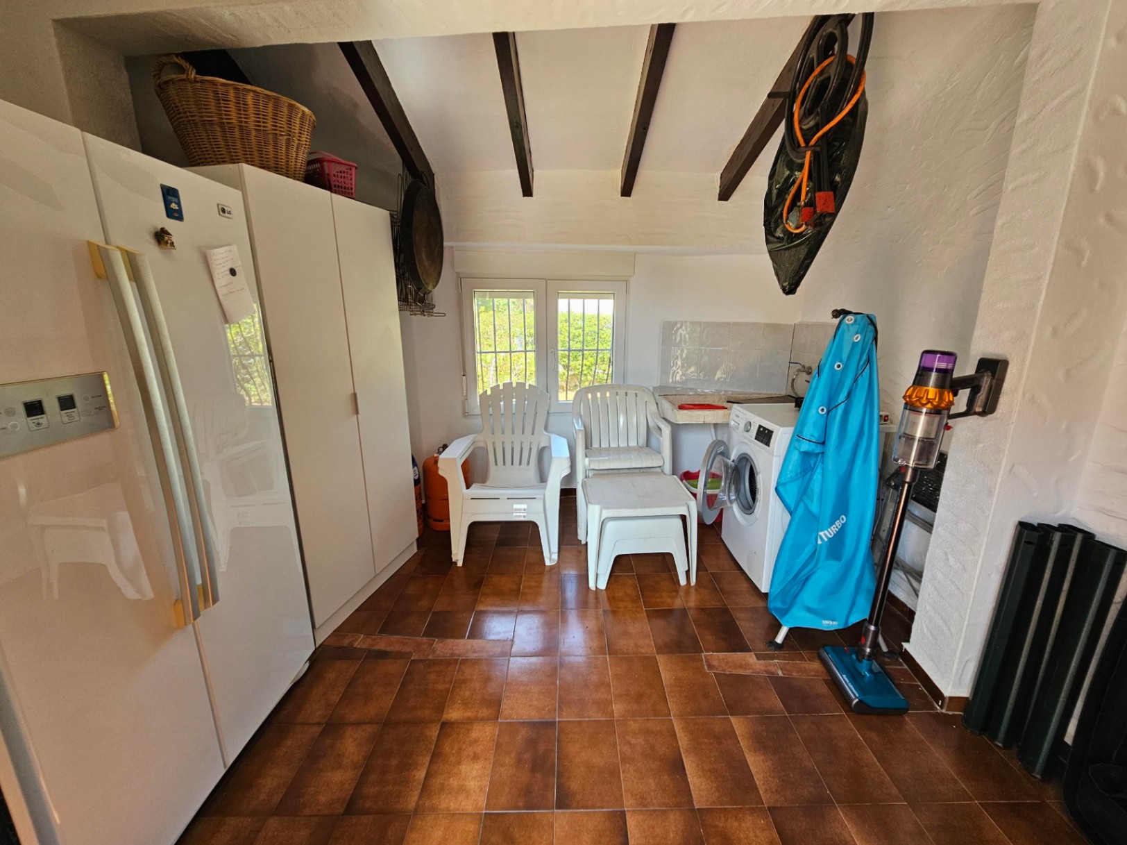 Espace buanderie avec sol en carreaux de terre cuite, poutres apparentes en bois sombre, lave-linge et armoires de rangement blanches.