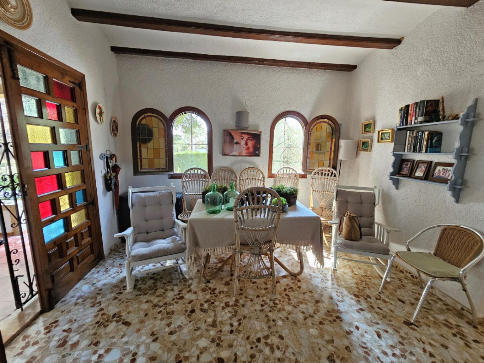 Espace repas avec sol en terrazzo, poutres en bois apparentes et fenêtres cintrées. Porte en bois à vitraux colorés et mobilier en rotin naturel.