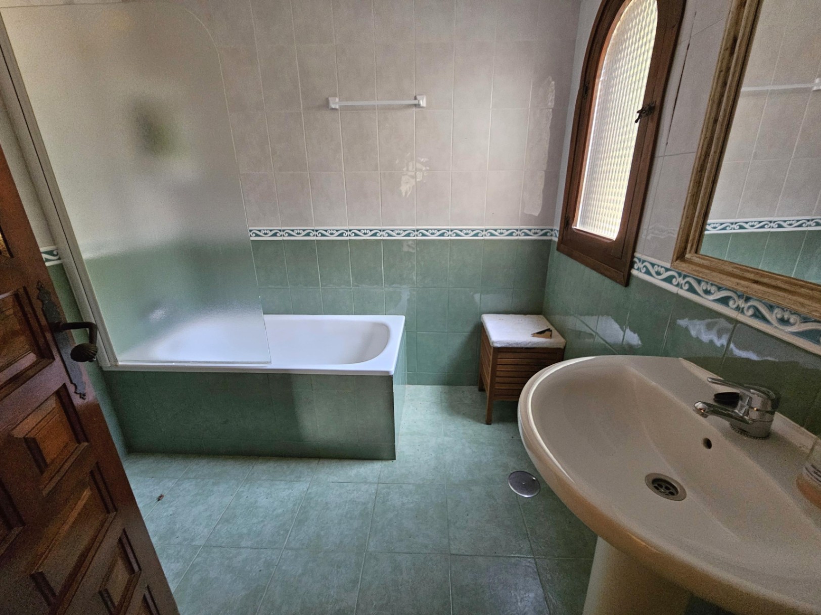 Salle de bain traditionnelle avec carrelage vert et blanc, baignoire à paroi en verre dépoli et lavabo sur colonne. Fenêtre cintrée en bois authentique.