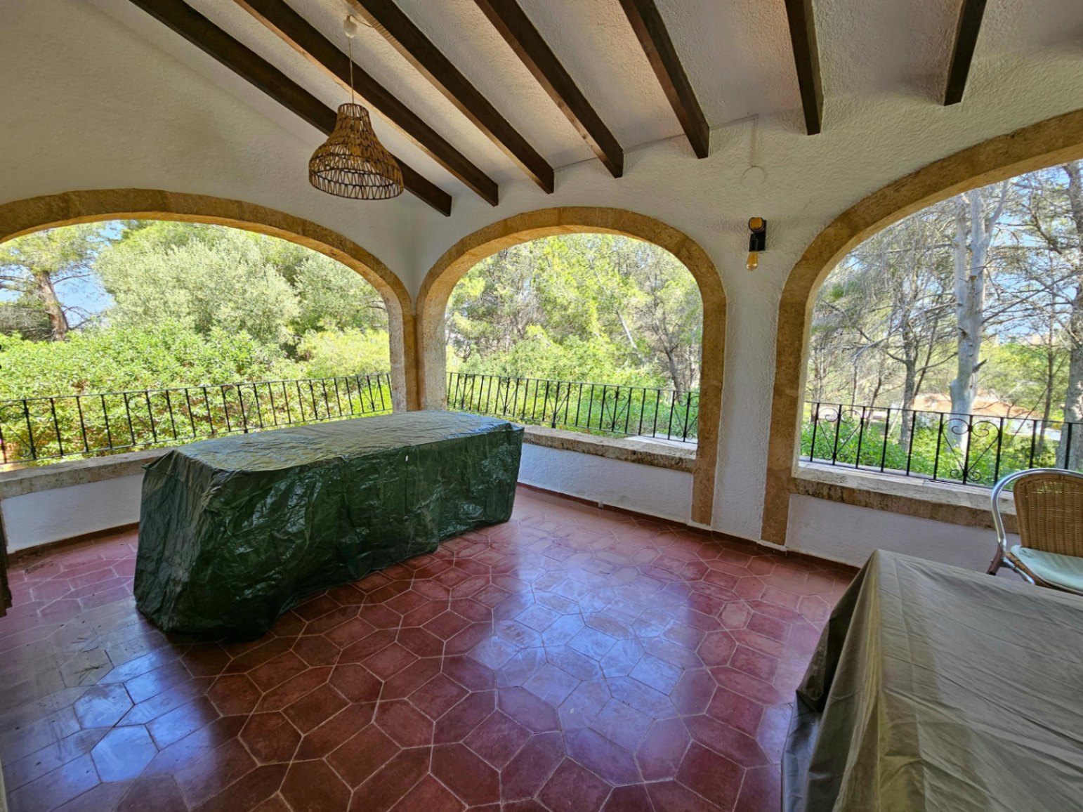 Terrasse couverte avec poutres en bois apparentes, sol en terre cuite hexagonale et arches en pierre donnant sur une zone boisée.