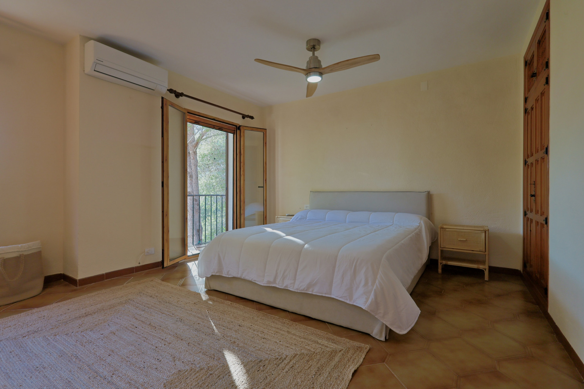 Chambre avec sol en terre cuite, lit double et ventilateur de plafond en bois. Portes-fenêtres ouvrant sur un balcon, placards intégrés en bois et climatisation murale.