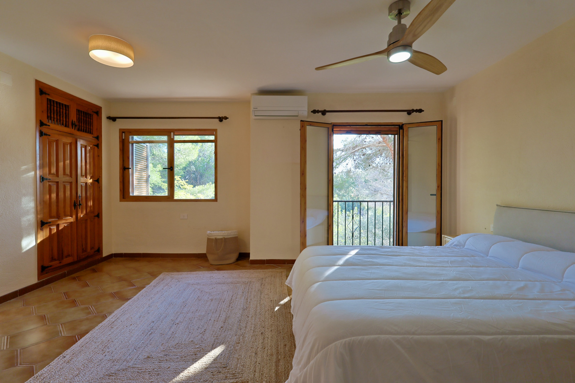 Chambre lumineuse avec sol en terre cuite, armoire encastrée en bois et balcon offrant une vue sur la verdure. Équipée d'un ventilateur de plafond et de la climatisation.