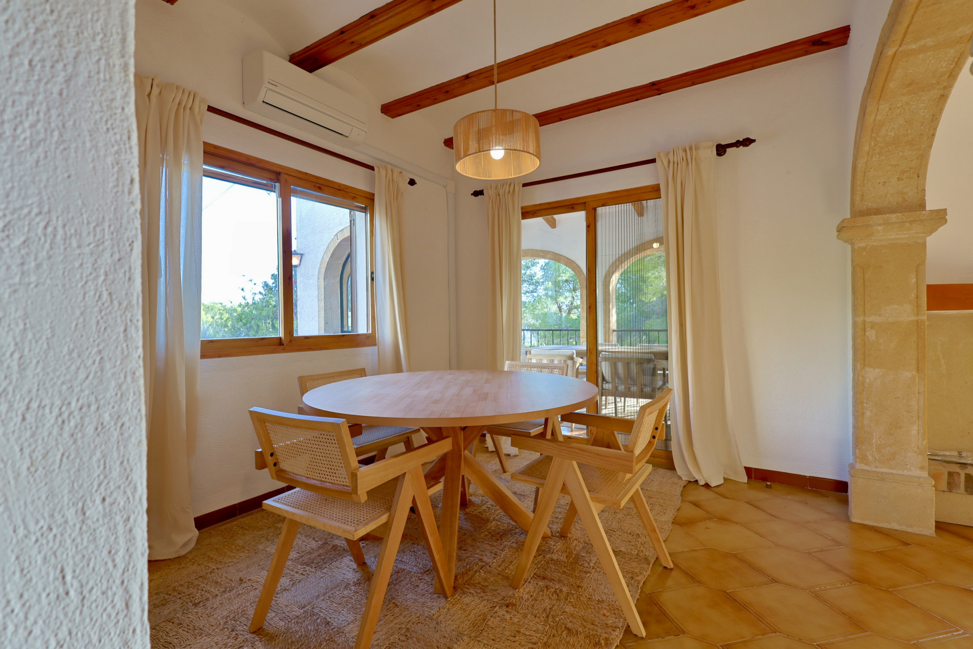 Coin repas avec table ronde en bois et chaises en cannage, sol en terre cuite, poutres apparentes et arche en pierre naturelle.