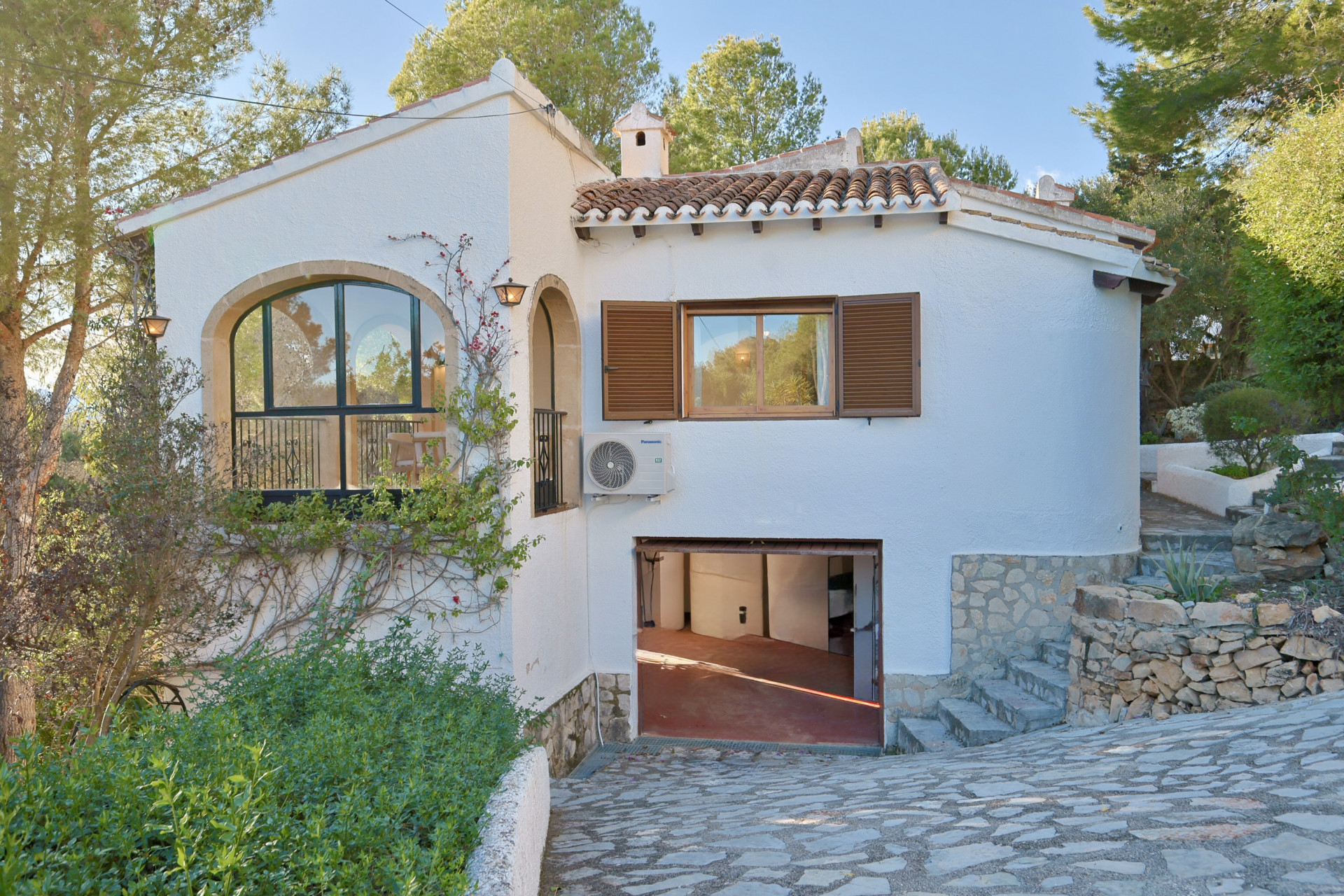 Façade d'une villa blanche avec arche en pierre, toiture en tuiles et garage en rez-de-chaussée, entourée d'une végétation méditerranéenne et de murets.