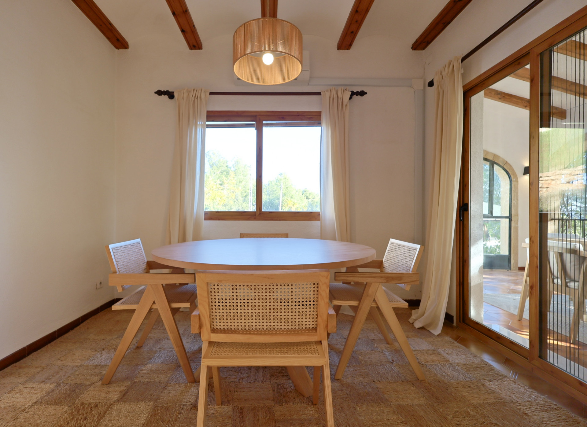 Salle à manger avec table ronde en bois, chaises en cannage et tapis en fibres naturelles, sous un plafond à poutres apparentes et suspension à lattes.