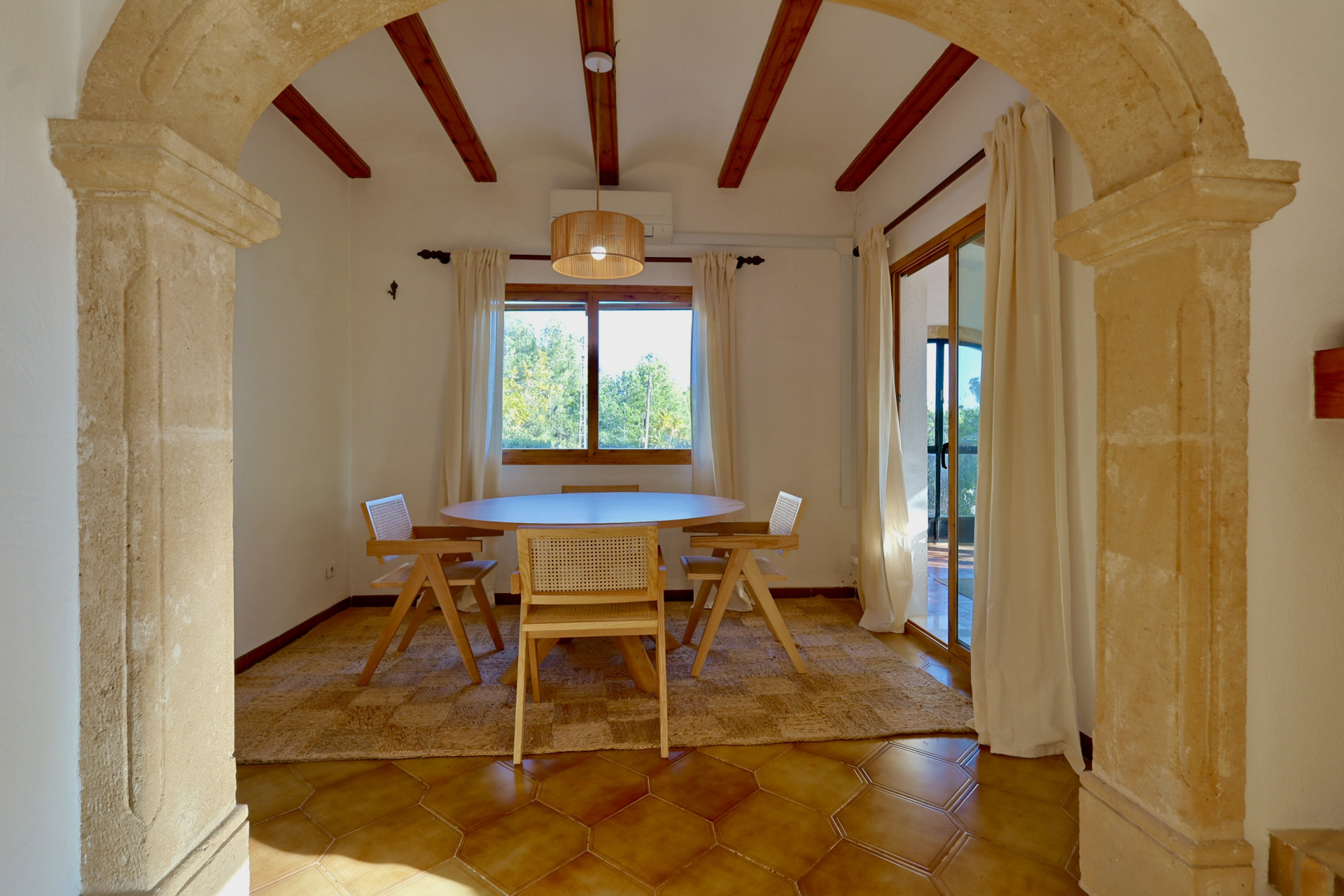 Salle à manger rustique encadrée par une arche en pierre, avec poutres en bois apparentes, sol en terre cuite hexagonale et mobilier en bois et rotin.
