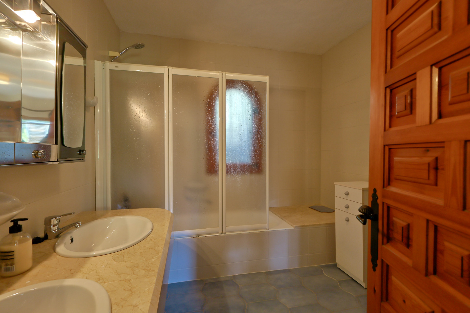Salle de bain avec double vasque en marbre, armoire de toilette miroir et baignoire avec paroi pliante dépolie. Carrelage bleu au sol et porte en bois massif.