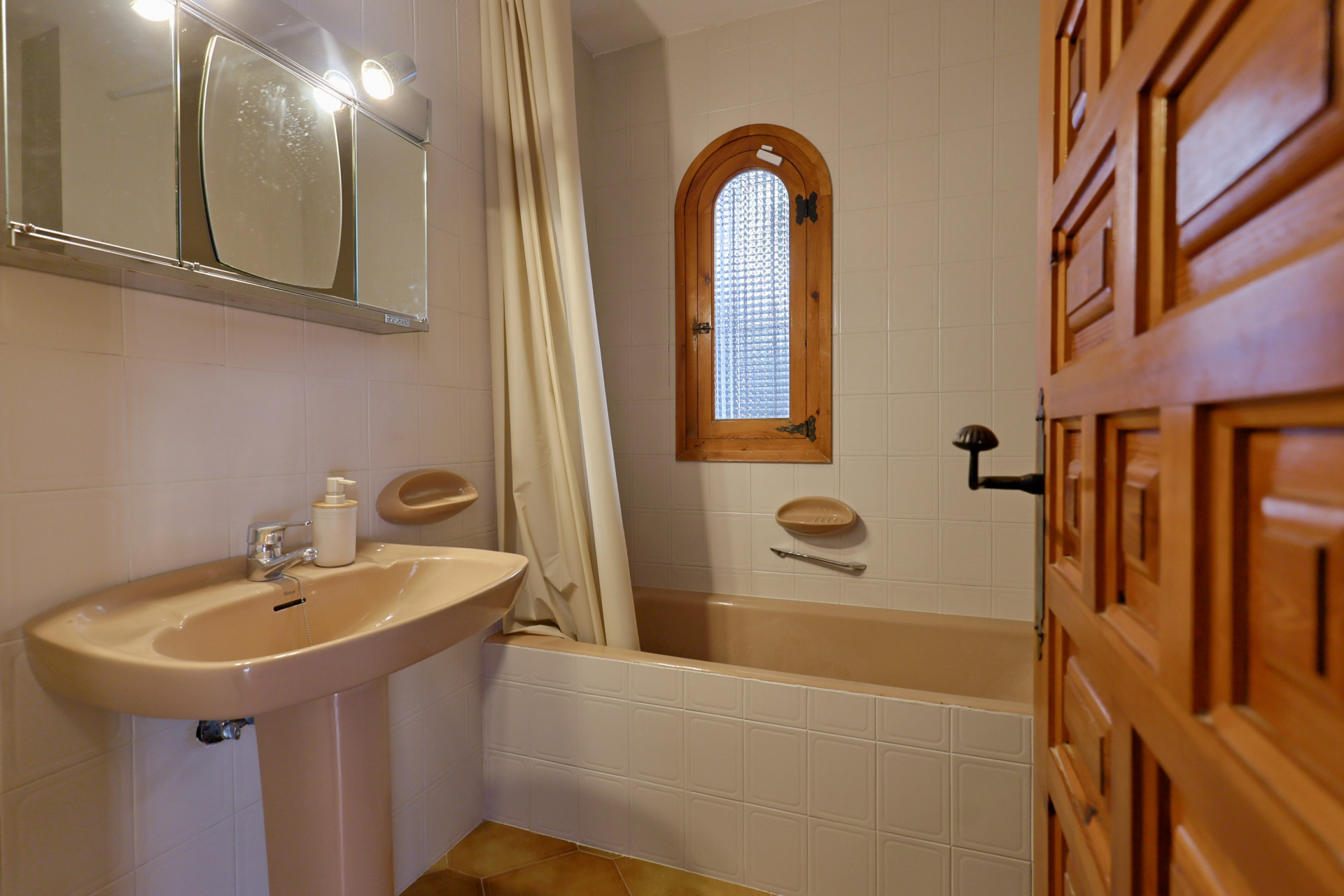 Salle de bain avec lavabo sur colonne et baignoire beige, carrelage mural blanc et fenêtre cintrée en bois avec verre dépoli.