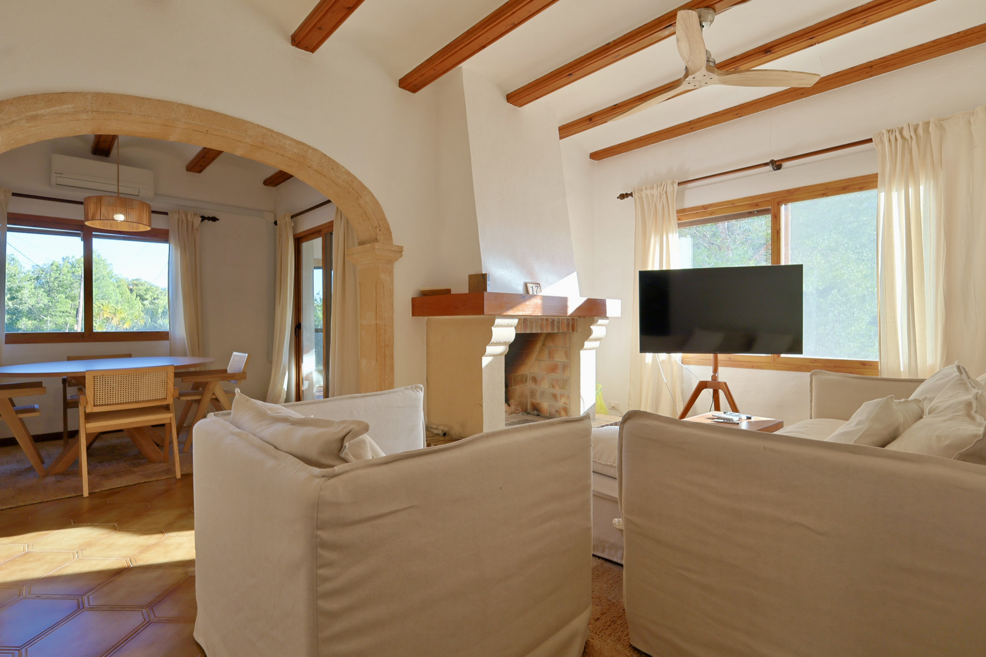 Salon avec poutres en bois apparentes, arche en pierre et cheminée en briques. Comprend des canapés blancs et un coin repas avec sol en terre cuite.