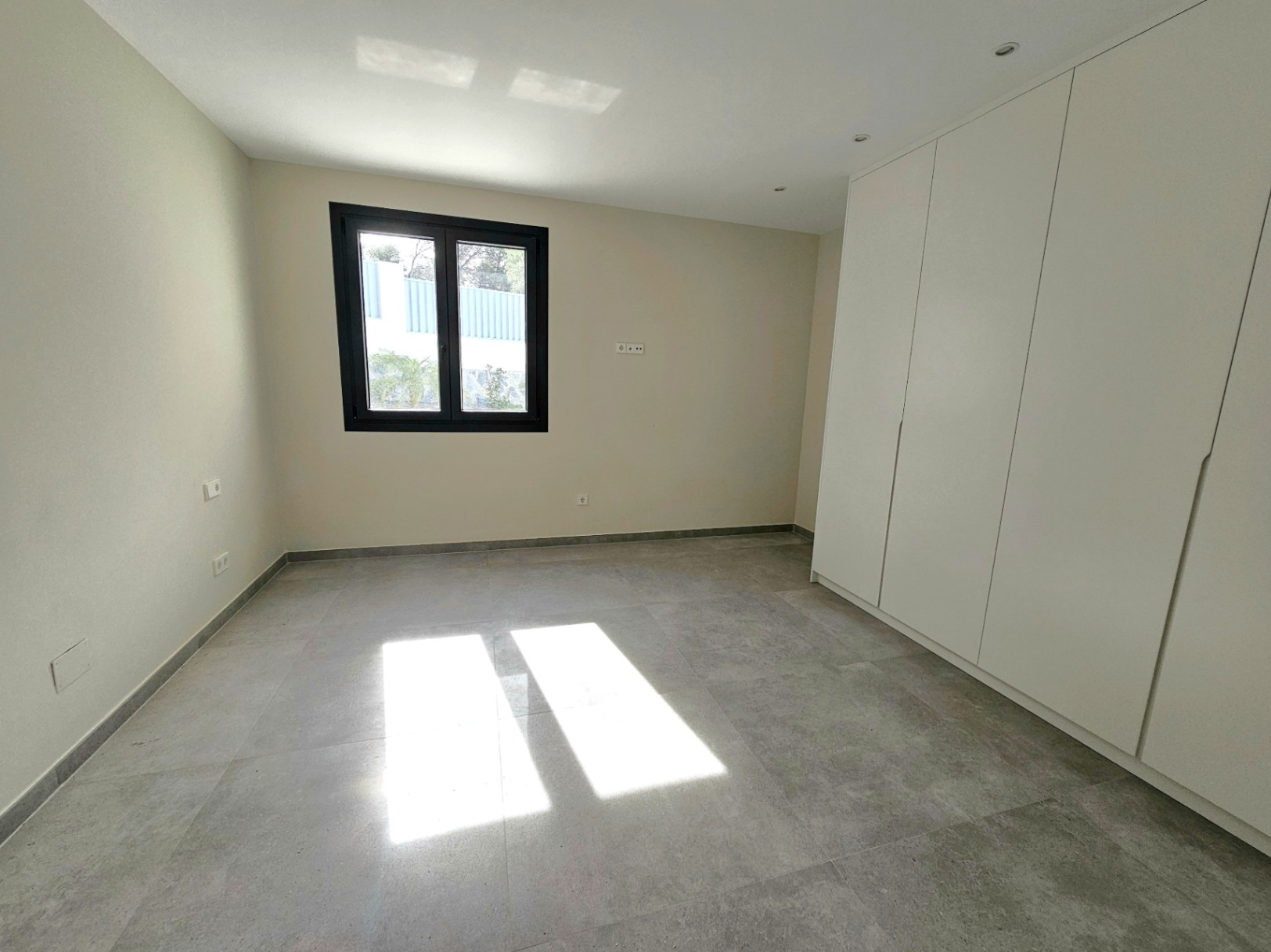 Chambre avec placard intégré blanc et sol en grès cérame gris, éclairée par la lumière naturelle de la fenêtre.