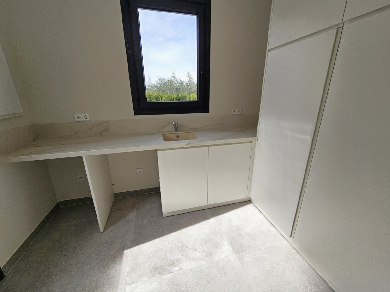Détail de la buanderie avec un comptoir en pierre claire, des armoires blanches et une fenêtre avec vue sur l'extérieur.
