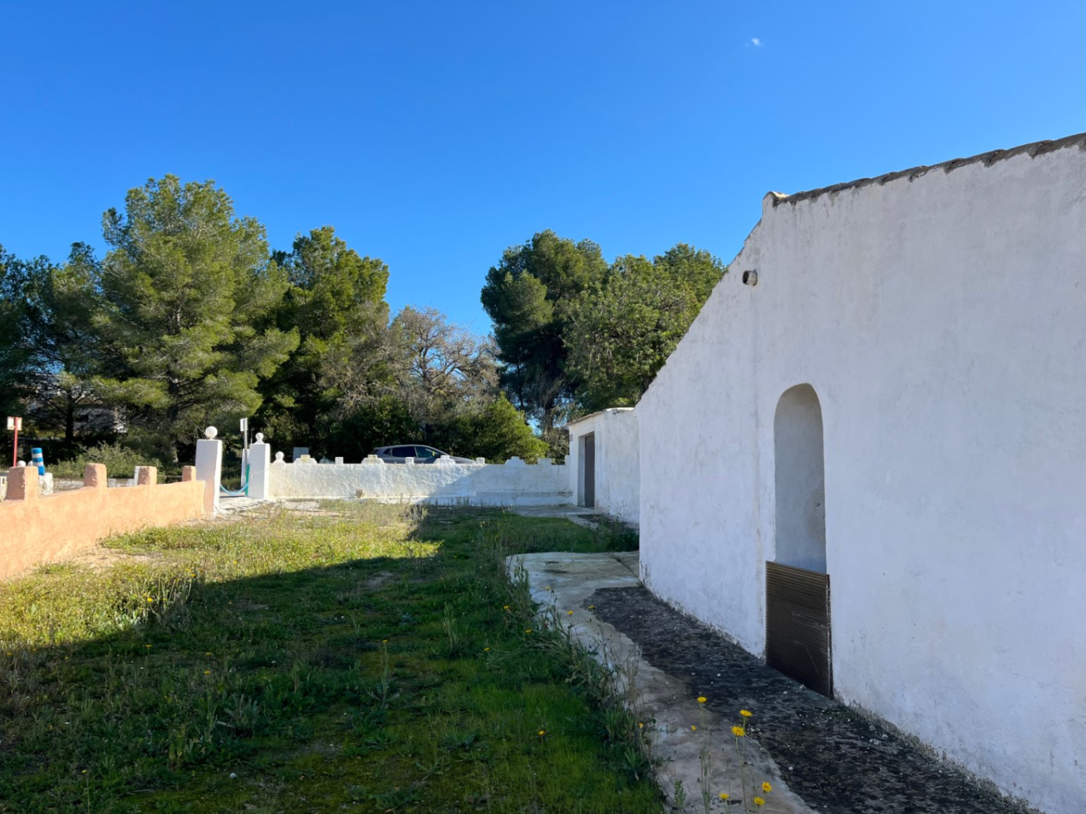Extérieur d'une finca traditionnelle à Jávea avec façade blanche, niche cintrée et terrain naturel entouré de pins sous un ciel bleu clair.