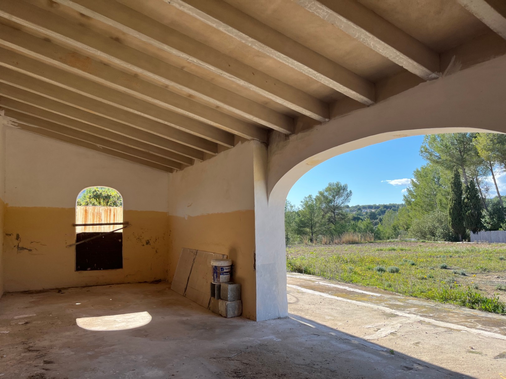 Porche à rénover avec plafond à poutres apparentes et une grande arche offrant une vue sur les champs environnants et les pins.